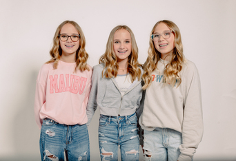 Three smiling teenage girls with long blonde hair wearing casual sweatshirts and ripped jeans