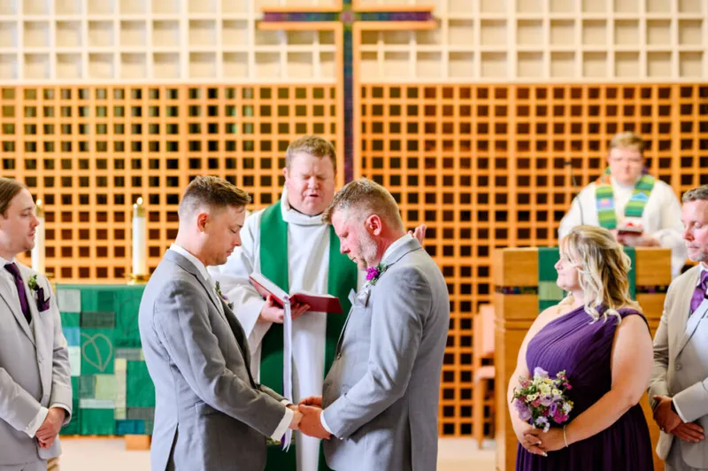 Two grooms in gray suits hold hands during a church wedding ceremony with an officiant in a green stole
