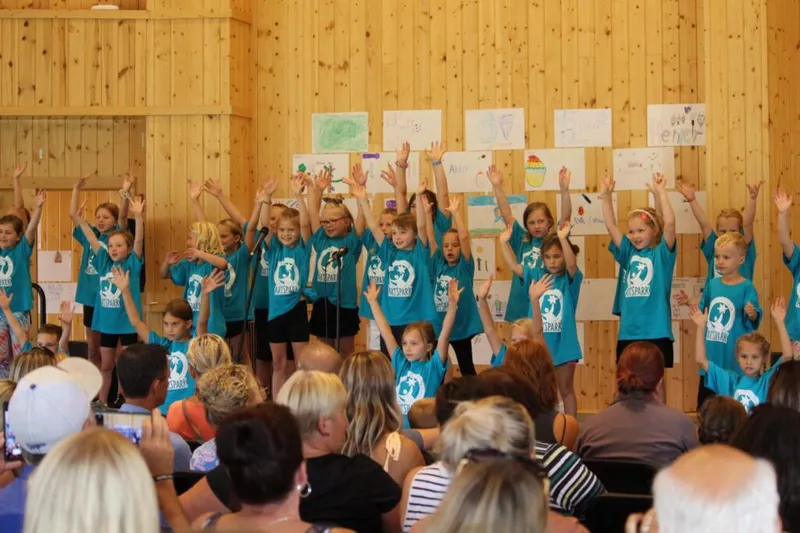 Children in matching teal T-shirts perform on stage with arms raised before a seated audience