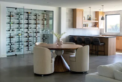 Round wood dining table with cream chairs beside a glass wine display wall and kitchen counter