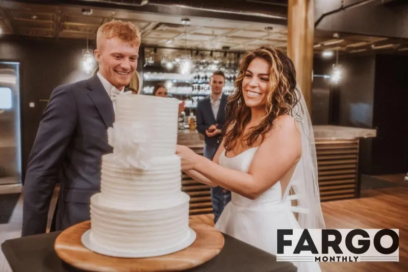 Smiling bride and groom cutting a white three-tier wedding cake in a dimly lit venue