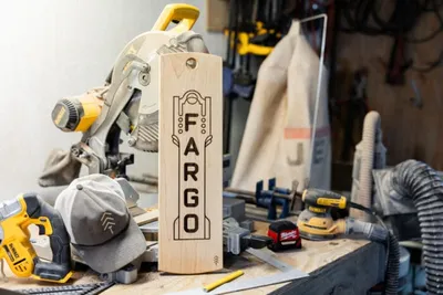 Wooden board engraved with 'Fargo' displayed on a workshop bench near a miter saw