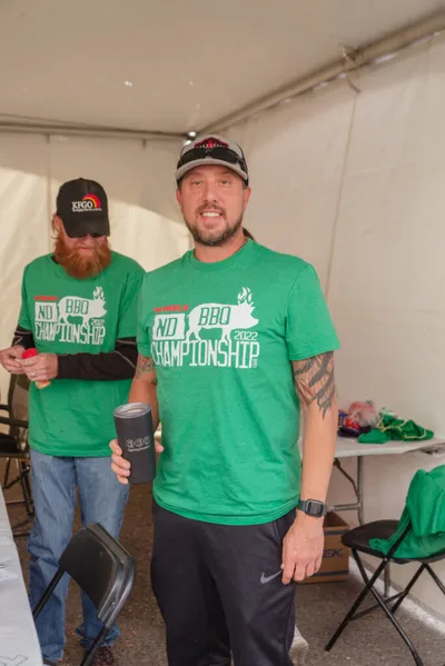 Two men in green ND BBQ Championship 2022 T-shirts under a tent, one holding a tumbler