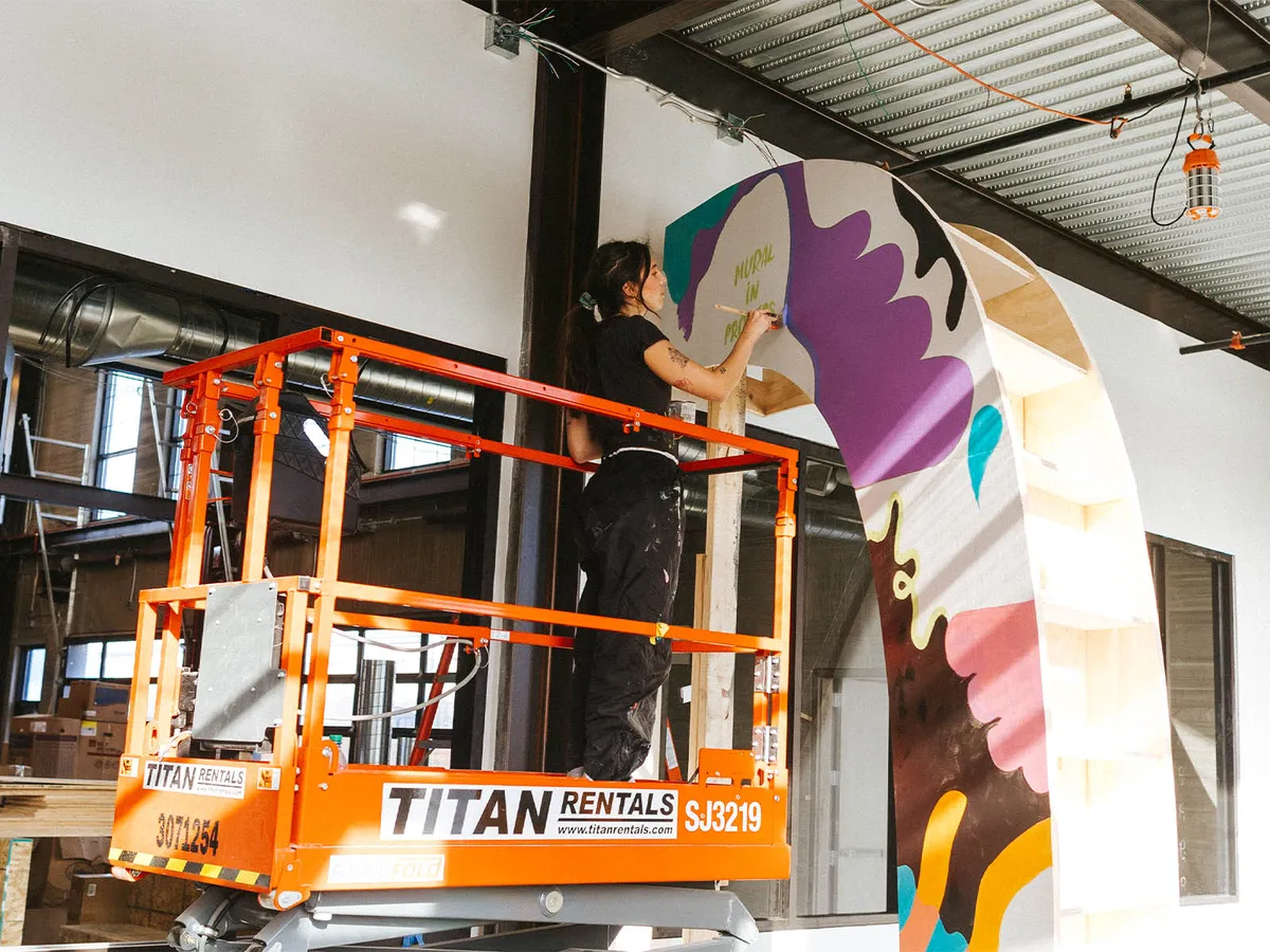 Artist on an orange scissor lift painting a colorful mural inside a building under construction