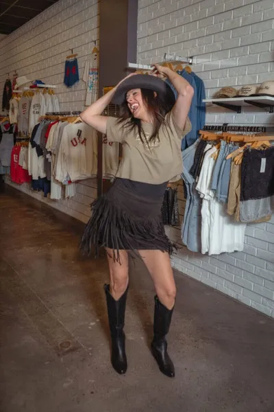 Woman in fringe skirt, cowboy boots, and wide-brim hat poses in a clothing boutique