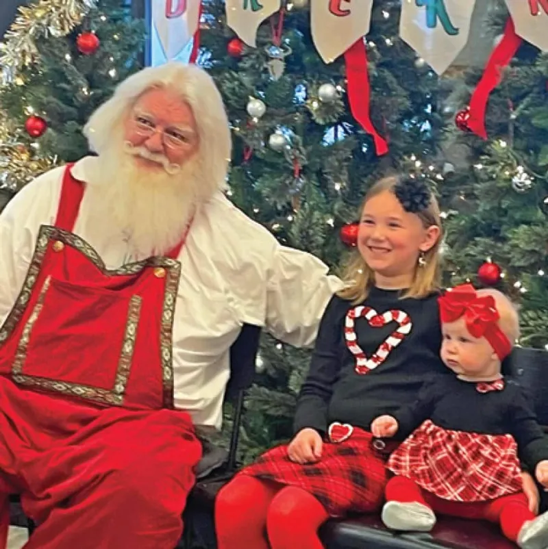 Santa Claus seated with a smiling young girl and a baby in front of a decorated Christmas tree