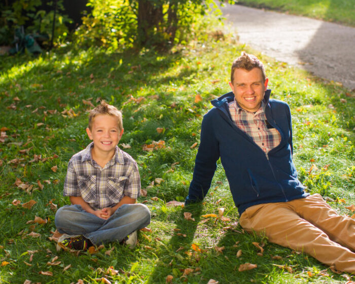 A smiling boy and a young man sit together on grass scattered with fallen leaves