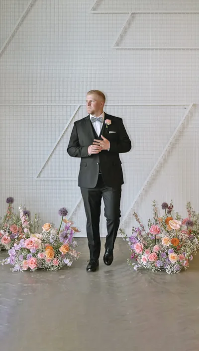 Groom in black tuxedo with pastel boutonniere stands before floral arrangements and white geometric backdrop
