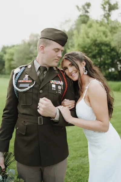 Bride in white gown leans smiling against groom in olive military dress uniform with medals
