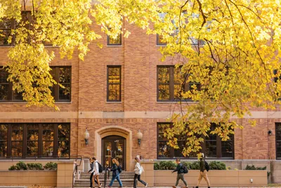 Students walking past a red brick campus building framed by bright yellow autumn foliage