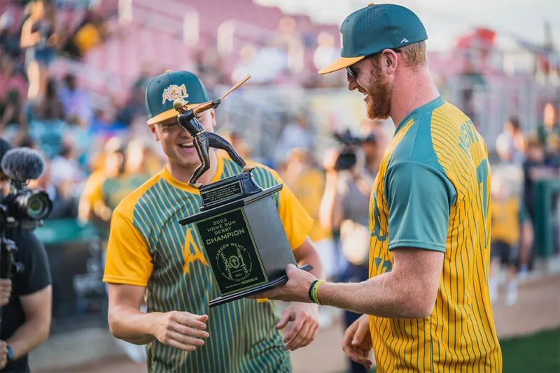 Two men in gold and teal striped jerseys share a 2022 Home Run Derby Champion trophy at a baseball stadium