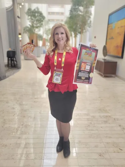 Smiling woman in red blazer holding a colorful game poster and cards in a bright atrium