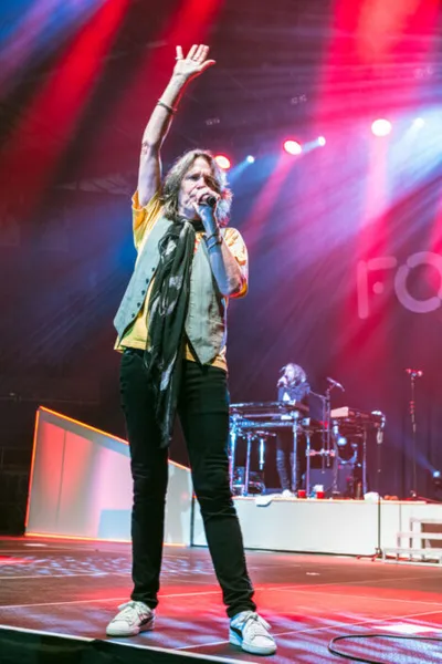 Long-haired male singer raises one arm while holding a mic on a red-lit concert stage