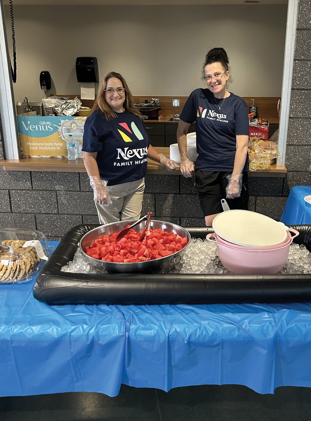 Two Nexus Family Healing team members in navy Nexus shirts standing behind a food table with a bowl of watermelon