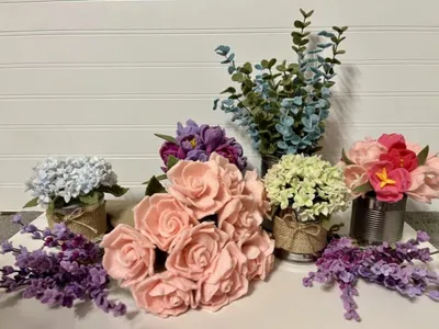 Felt and artificial flower arrangements in burlap-wrapped tins with loose purple stems on a white surface