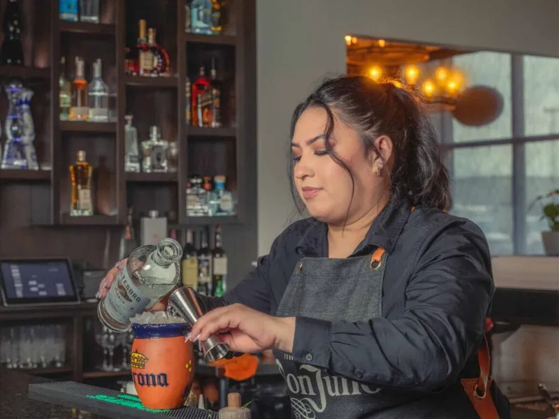 Bartender in apron pouring spirits into a jigger over an orange mule mug at a bar