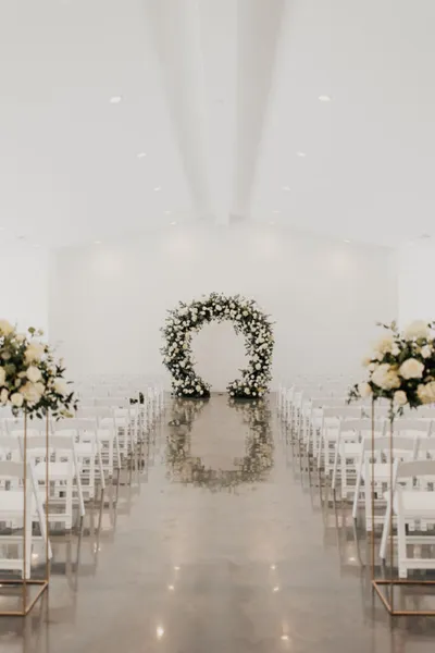 White wedding ceremony venue with floral arch at the end of a polished aisle lined with white chairs and tall floral