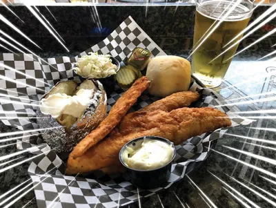 Fried fish strips in a basket with baked potato, coleslaw, roll, and dipping sauce beside a beer
