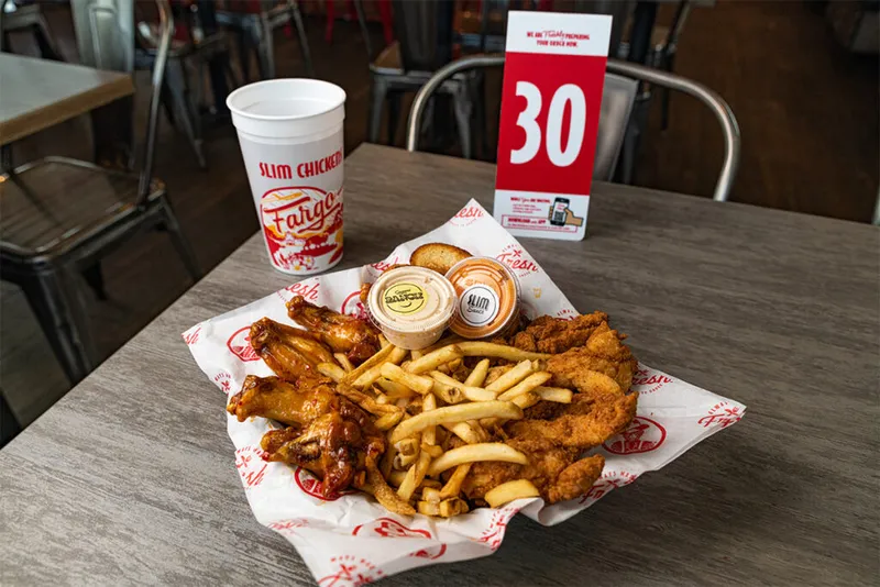 Basket of chicken tenders, wings, and fries with dipping sauces and a branded cup at Slim Chickens