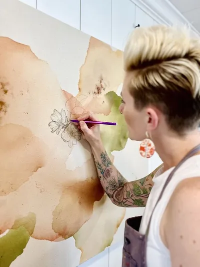 Tattooed artist with short blonde hair drawing a flower on a large watercolor mural