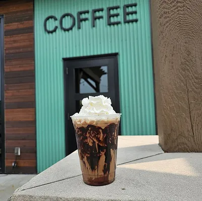 Iced mocha topped with whipped cream in front of a teal corrugated metal building with COFFEE sign
