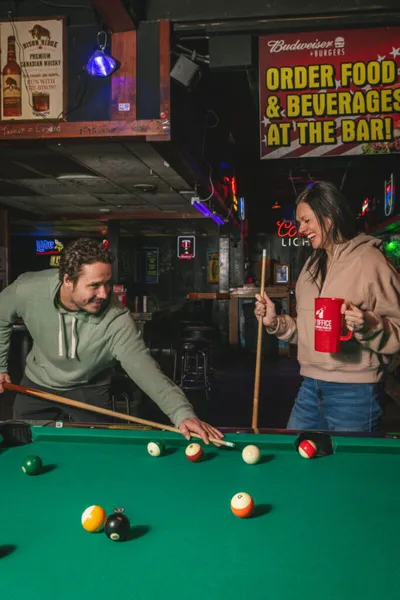 Man lining up a pool shot while a woman holds a red mug at a dimly lit bar