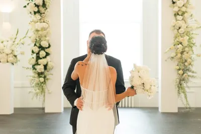 Bride and groom embracing from behind amid tall white floral arrangements
