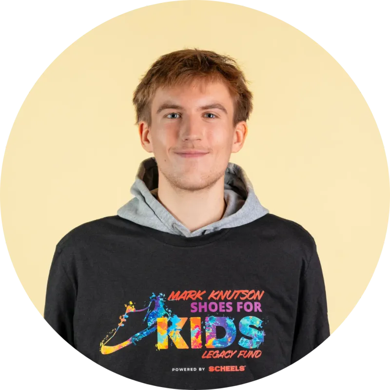 Young man smiling in a black Mark Knutson Shoes for Kids Legacy Fund T-shirt over a gray hoodie