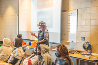 A masked presenter gestures toward a projected slide before a classroom of masked students