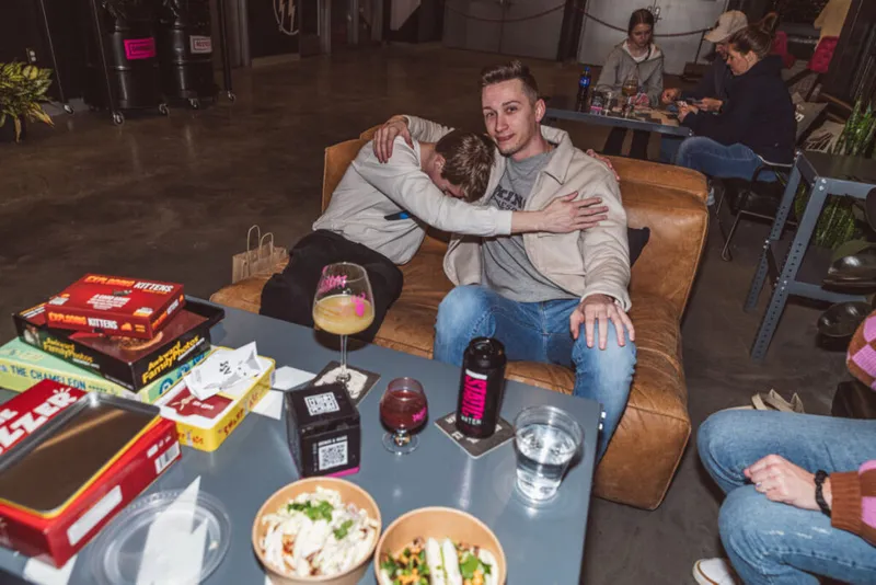Two friends hugging on a leather sofa surrounded by board games, cocktails, and food bowls on a low table