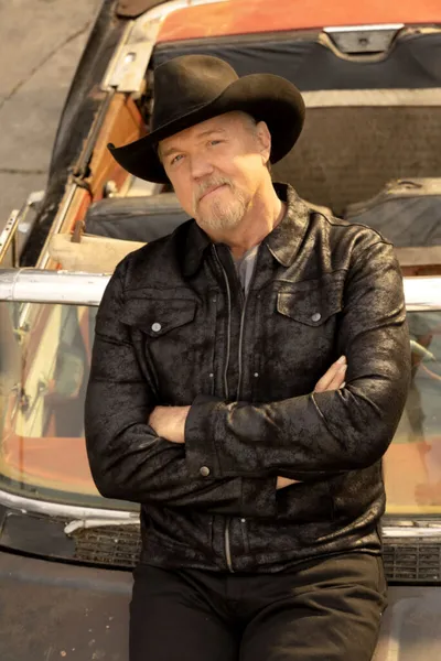 Man in black cowboy hat and dark jacket leaning against a vintage car with arms crossed