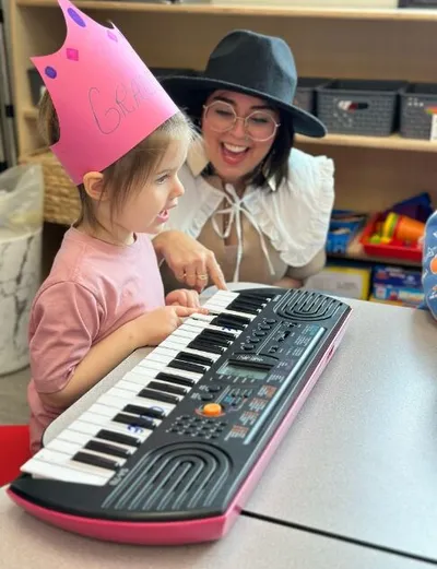 Young girl in a paper crown plays a small keyboard while a smiling woman in a wide-brim hat guides her