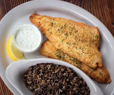 Golden pan-fried fish fillet with herbs, lemon wedge, tartar sauce, and wild rice on a white plate