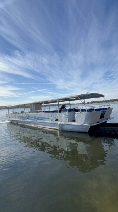 Large white pontoon boat with open-air canopy docked on a calm lake under a wispy blue sky