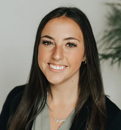 Smiling young woman with long dark hair wearing a sage green top and black blazer