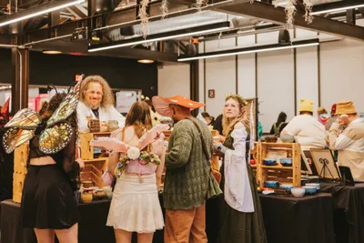 Costumed vendors and visitors mingle around wooden display tables at an indoor craft fair