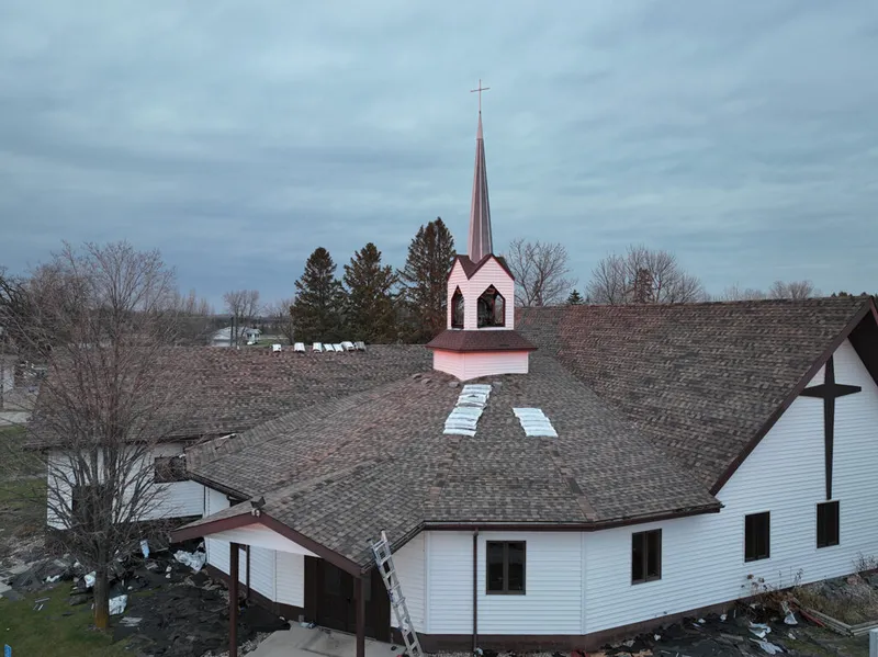 Aerial view of a white church with a red steeple mid-roofing repair under overcast skies