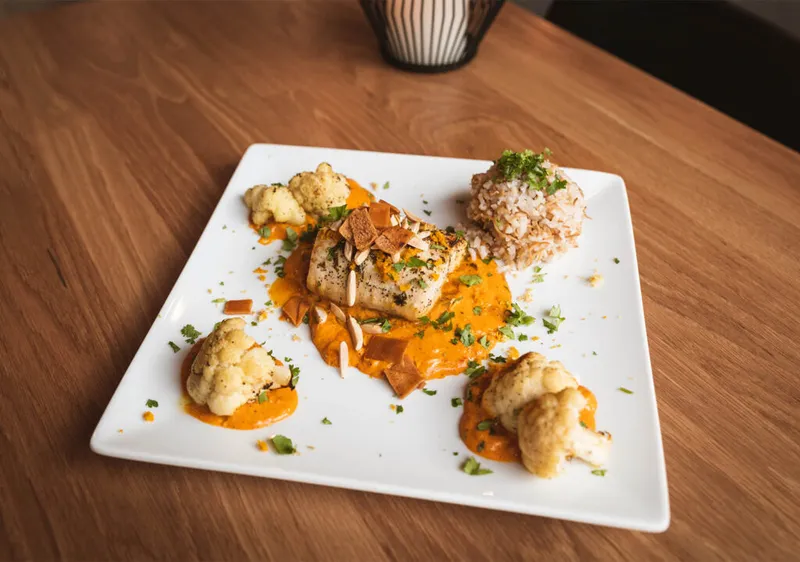 Plated white fish over orange sauce with roasted cauliflower, sliced almonds, and herbed rice on a square white plate