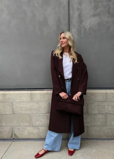 Blonde woman in a long dark burgundy coat, white tee, wide-leg jeans, and red flats
