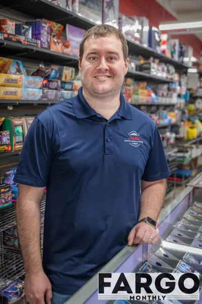 Smiling man in a navy polo standing inside a hobby shop with shelves of trading cards and games