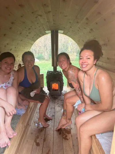 Four women in swimwear sitting on wooden benches inside a barrel sauna with a wood-burning stove