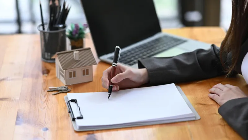 Person signing document on clipboard beside a small house model and keys on a wooden desk