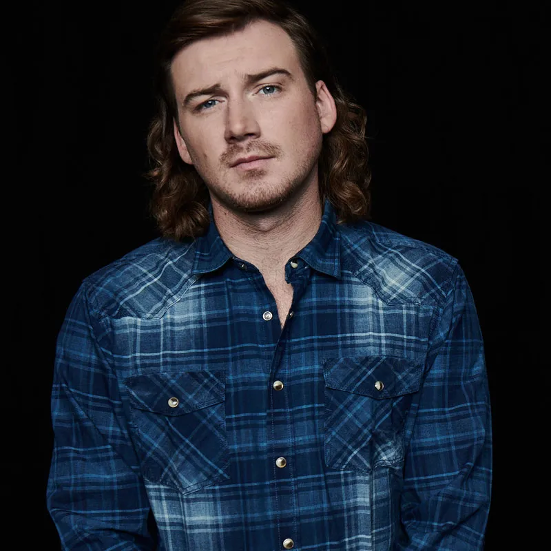 Young man with shoulder-length brown hair in a blue plaid western snap shirt against a dark background