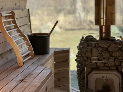 Wood-fired sauna stove loaded with stones beside a curved backrest and black bucket on a pine bench