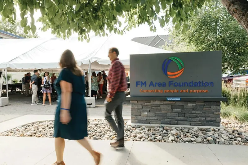Two people walk past the FM Area Foundation monument sign at an outdoor event with a tent and crowd behind