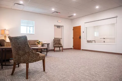 Carpeted funeral home waiting room with patterned armchairs, a small table, and a wood-framed service window