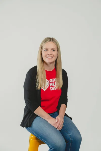 Smiling woman in a red 'Giving Heart Day' T-shirt and black cardigan seated on a yellow stool