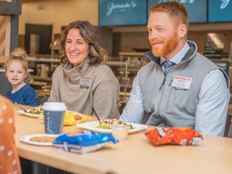 Two smiling Scheels employees wearing name badges sit at a dining table with food, a young redheaded girl nearby