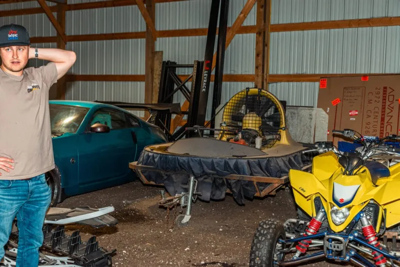 Young man in a cap stands in a metal warehouse beside a teal car, hovercraft, and yellow ATV