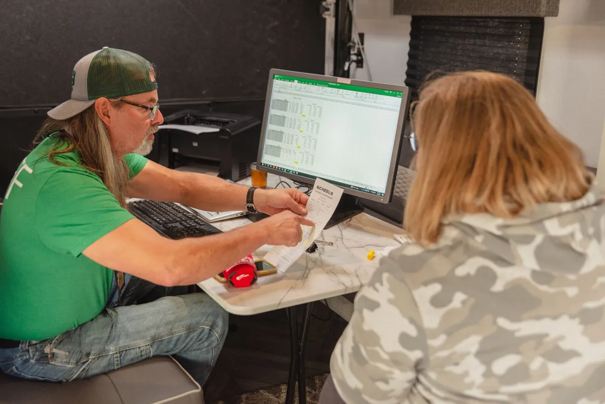Man in green cap reviews a receipt with a woman beside a spreadsheet on a desktop monitor